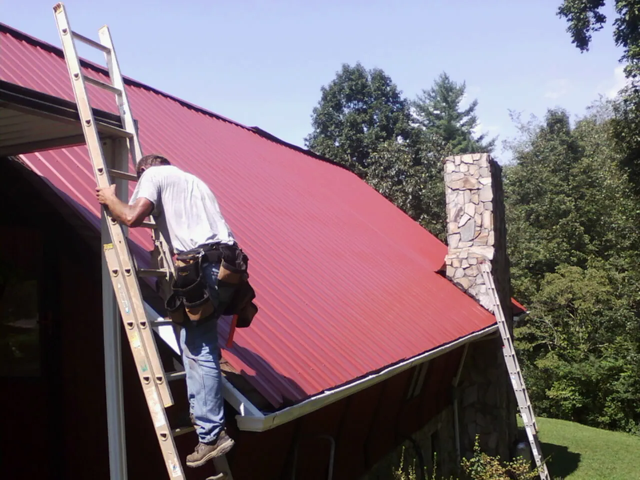 Roofer on a ladder beside a red metal roof and stone chimney — Soffit & Fascia Repair by Roofing Brothers in Twain